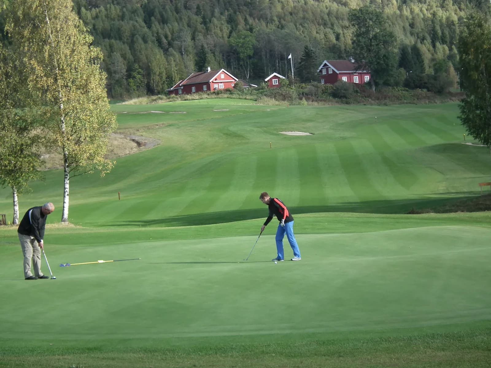 Two golfers on the putting green on hole 8 at Aas Gaard Golf Park with red houses and forest